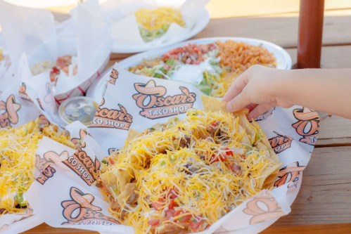 A hand reaching for a loaded nacho platter topped with cheese, alongside colorful sides from Oscar's Taco Shop.