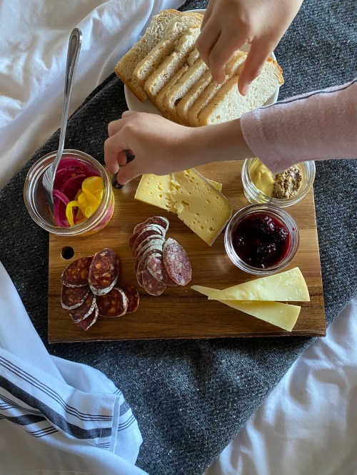 A wooden board with assorted cheeses, cured meats, pickled vegetables, mustard, and jam, with a hand reaching for food.