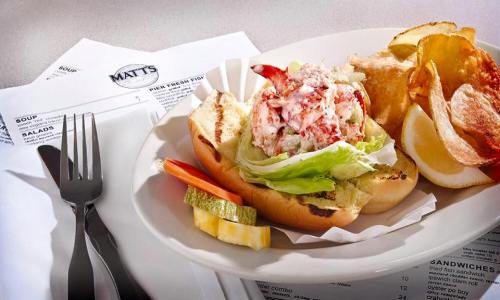 A lobster roll on a toasted bun with lettuce, served with potato chips and lemon wedges on a plate.