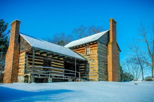 A rustic log cabin with a snow-covered roof, surrounded by a winter landscape and clear blue sky.