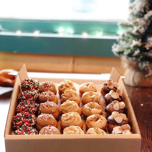 A box of assorted decorated donuts, featuring festive toppings and designs, displayed near a small Christmas tree.