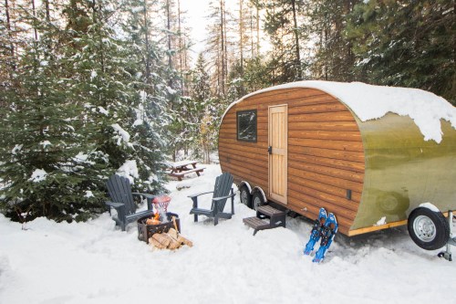 A cozy trailer in a snowy forest, with a fire pit and chairs nearby, surrounded by tall trees.