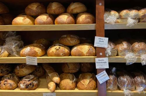 A display of various artisanal breads on wooden shelves, including sourdough, jalapeño cheddar, and olive herb loaves.