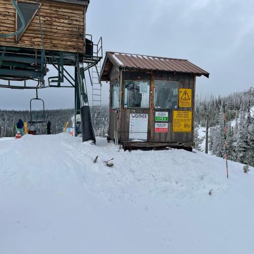 A small wooden weather station near a ski lift, surrounded by snow and trees, with various signs posted on it.