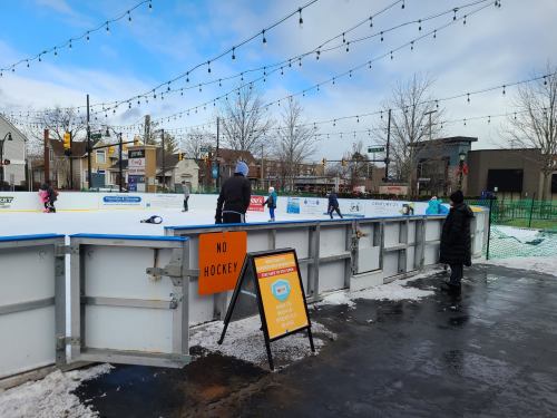Outdoor ice skating rink with people skating, a "No Hockey" sign, and festive lights strung above. Snow on the ground.