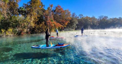 Three people paddleboarding on a misty, clear lake surrounded by colorful autumn trees.