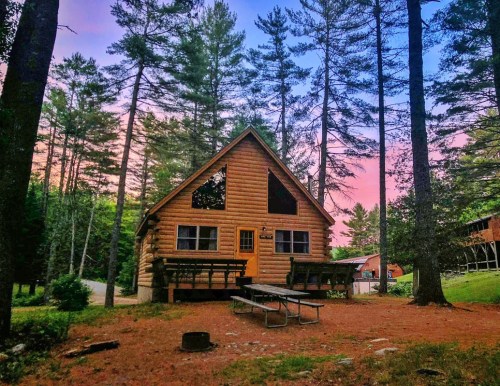 A cozy log cabin surrounded by tall trees, with a pink and blue sky at sunset. Picnic tables are in front.