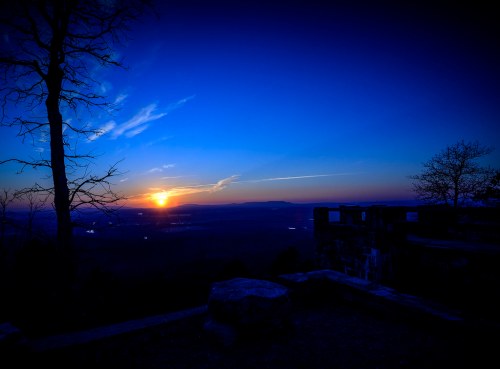 A serene sunset over a valley, with a silhouette of trees and a stone structure in the foreground.