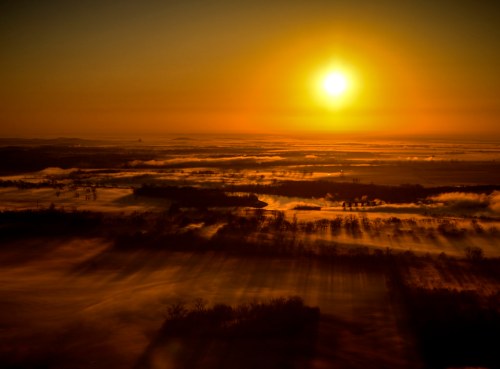 A golden sunrise casts long shadows over a misty landscape, with rolling hills and trees silhouetted against the sky.
