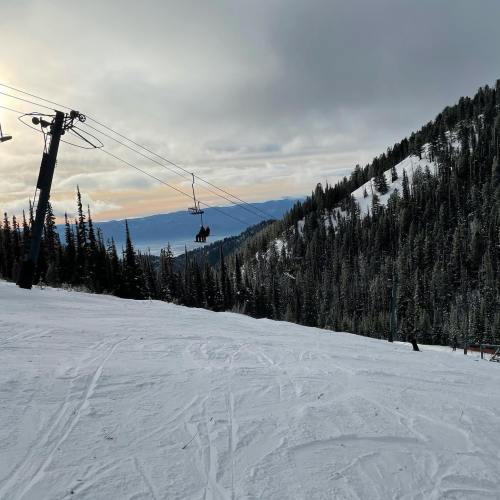 A snowy mountain landscape with a ski lift and a skier in the foreground, surrounded by evergreen trees.
