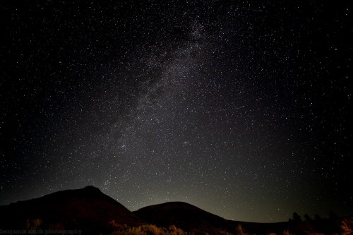 A starry night sky over dark hills, with the Milky Way visible and a hint of light on the horizon.