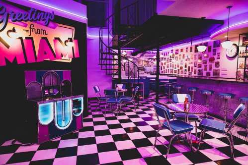 A retro diner with a black and white checkered floor, neon lights, and a "Greetings from Miami" sign on the wall.