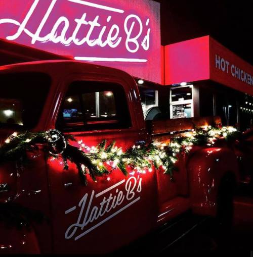 A vintage red truck decorated with lights and greenery in front of Hattie B's Hot Chicken restaurant at night.