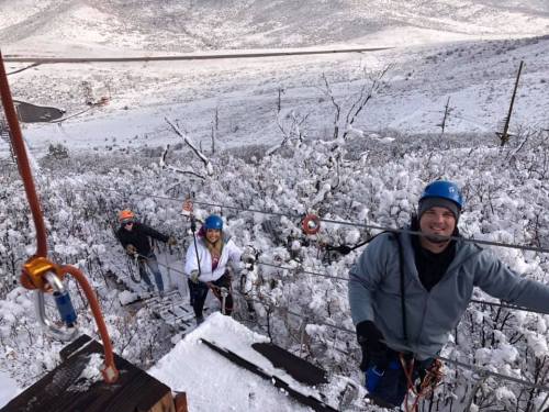 Three climbers in winter gear ascend a snowy, mountainous landscape, with trees covered in snow and a road visible below.