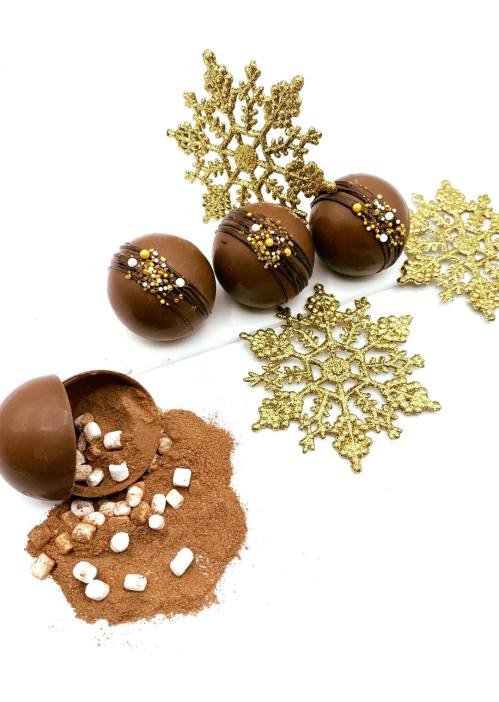 Three chocolate spheres with decorations, one open revealing cocoa and marshmallows, surrounded by gold snowflakes.