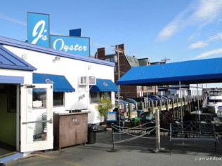 Exterior of J's Oyster restaurant with blue awning, outdoor seating, and a view of parked cars and the waterfront.