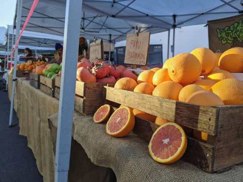 A market stall displaying wooden crates filled with oranges, persimmons, and other fruits under white tents.