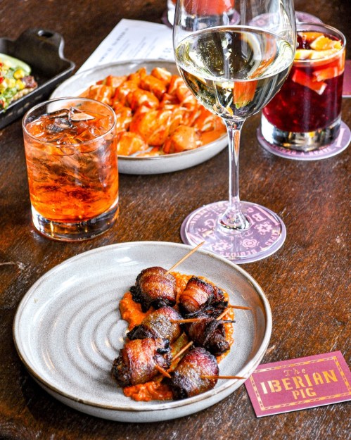 A plate of bacon-wrapped appetizers on a table with drinks and other dishes in the background.