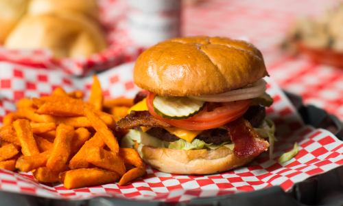 A juicy burger with bacon, cheese, lettuce, tomato, and pickles, served with a side of sweet potato fries on a checkered tray.