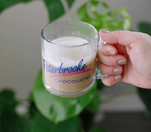 A hand holding a glass mug with coffee, featuring the logo "Peterbrooke Chocolatier," against a green plant background.