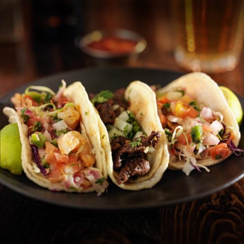 Three tacos on a black plate, filled with shrimp, beef, and fresh toppings, served with lime wedges and salsa.