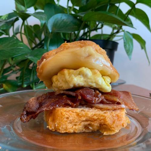 A close-up of a sandwich with bacon, scrambled eggs, and a biscuit, set against a backdrop of green plants.
