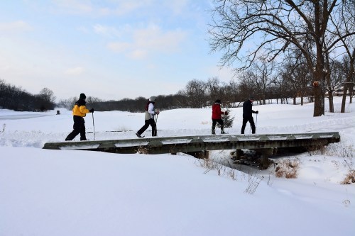 Four people snowshoeing across a wooden bridge in a snowy landscape with trees in the background.