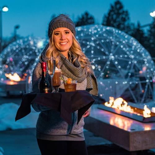 A smiling woman holds a tray with drinks, standing near illuminated igloos and fire pits in a winter setting.