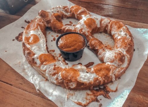 A large, soft pretzel topped with cheese and served with a side of dipping sauce on a paper-lined plate.