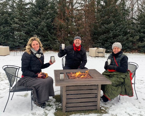 Three people sitting around a fire pit in the snow, holding mugs and wearing warm clothing, with trees in the background.