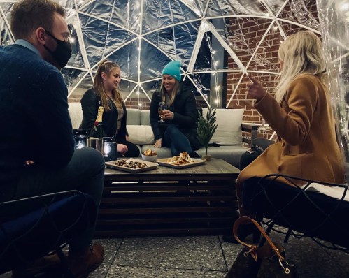 Group of four people in a cozy outdoor igloo, enjoying food and drinks, wearing masks and winter attire.