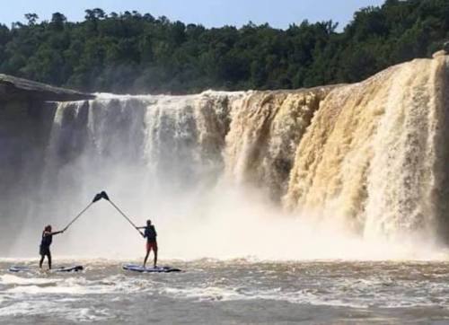 Two people paddleboarding near a large waterfall, surrounded by lush greenery and mist from the cascading water.
