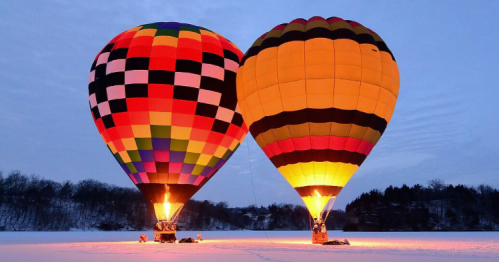 Two colorful hot air balloons are illuminated against a twilight sky, set in a snowy landscape.