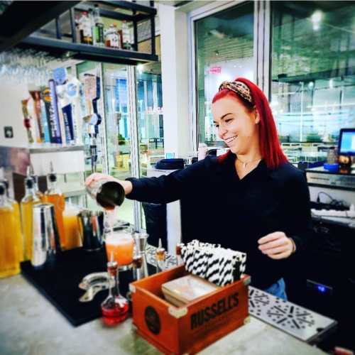 A bartender with red hair mixes a cocktail at a modern bar, smiling as she pours from a shaker.