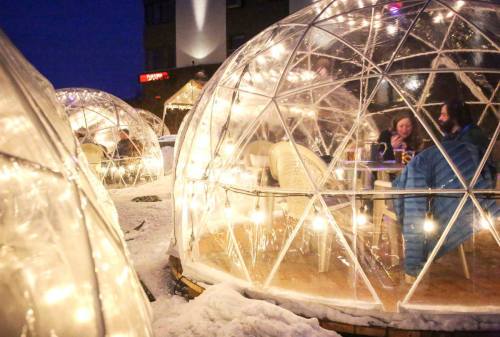 Cozy outdoor dining igloos illuminated with string lights, surrounded by snow and a darkening sky.