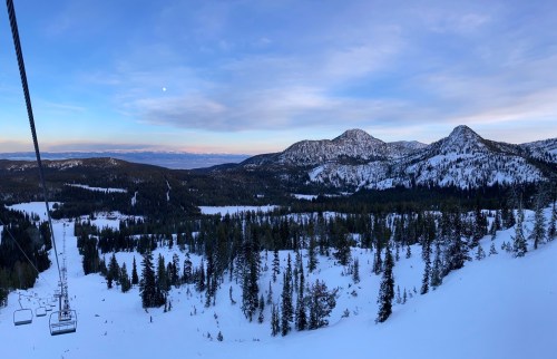 A scenic view from a ski lift, showcasing snow-covered mountains and a forested valley under a clear sky.