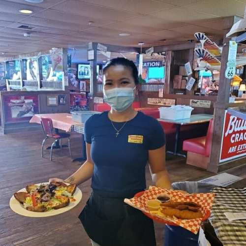 A waitress in a mask holds two plates of food in a restaurant with a casual atmosphere.