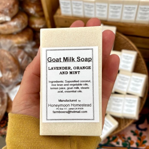 A hand holding a bar of goat milk soap with a label listing ingredients: lavender, orange, mint, and more.
