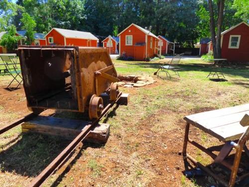 Rusty mining cart on tracks in a grassy area with red cabins and chairs in the background.