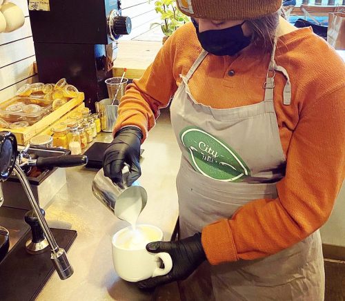 A barista in a mask and gloves pours steamed milk into a cup at a coffee shop.