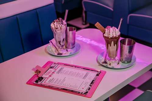 Two milkshakes with whipped cream and toppings sit on a table with a menu in a retro diner setting.