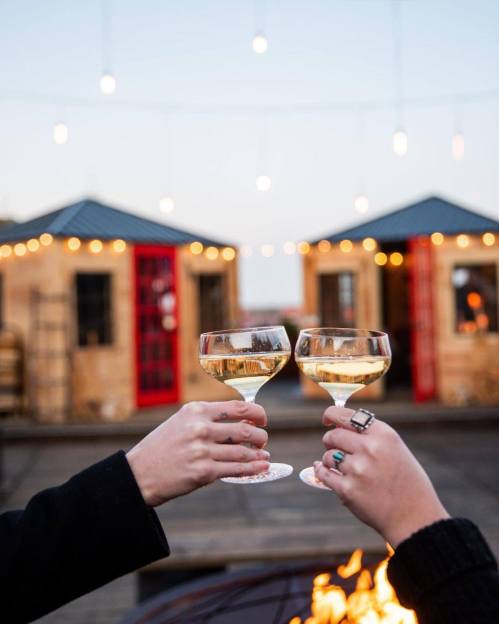 Two hands clink champagne glasses in front of cozy cabins, with string lights glowing in the evening sky.