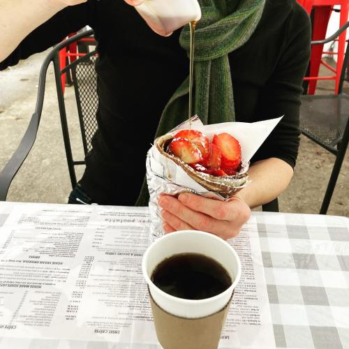 A person drizzles syrup over a crepe filled with strawberries, next to a cup of coffee on a checkered tablecloth.