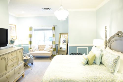 A cozy bedroom with a large bed, decorative pillows, a stylish armchair, and soft blue walls. Natural light from a window.