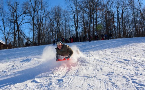 A person sleds down a snowy hill, surrounded by trees and a crowd of people in the background enjoying winter activities.
