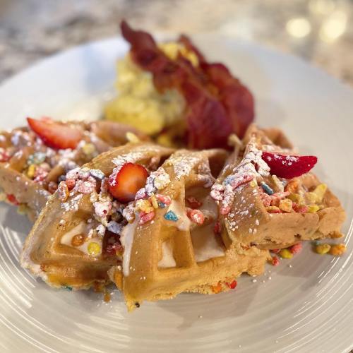 A plate of waffles topped with strawberries, colorful cereal, and powdered sugar, accompanied by crispy bacon.