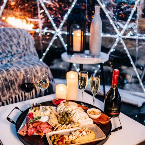 A cozy indoor setting with a charcuterie board, champagne, wine glasses, and candles, surrounded by twinkling lights.