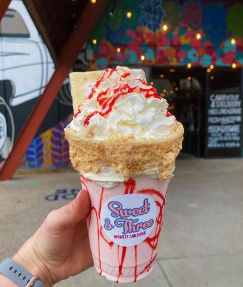 A hand holds a large milkshake topped with whipped cream, graham cracker crumbs, and a drizzle of red syrup.