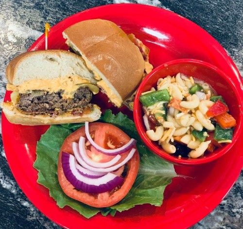 A red plate with a cheeseburger, lettuce, tomato, onion, and a side of pasta salad with vegetables.