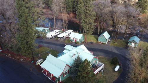 Aerial view of a quaint lodge with green buildings, RVs, and a river surrounded by trees in a serene landscape.